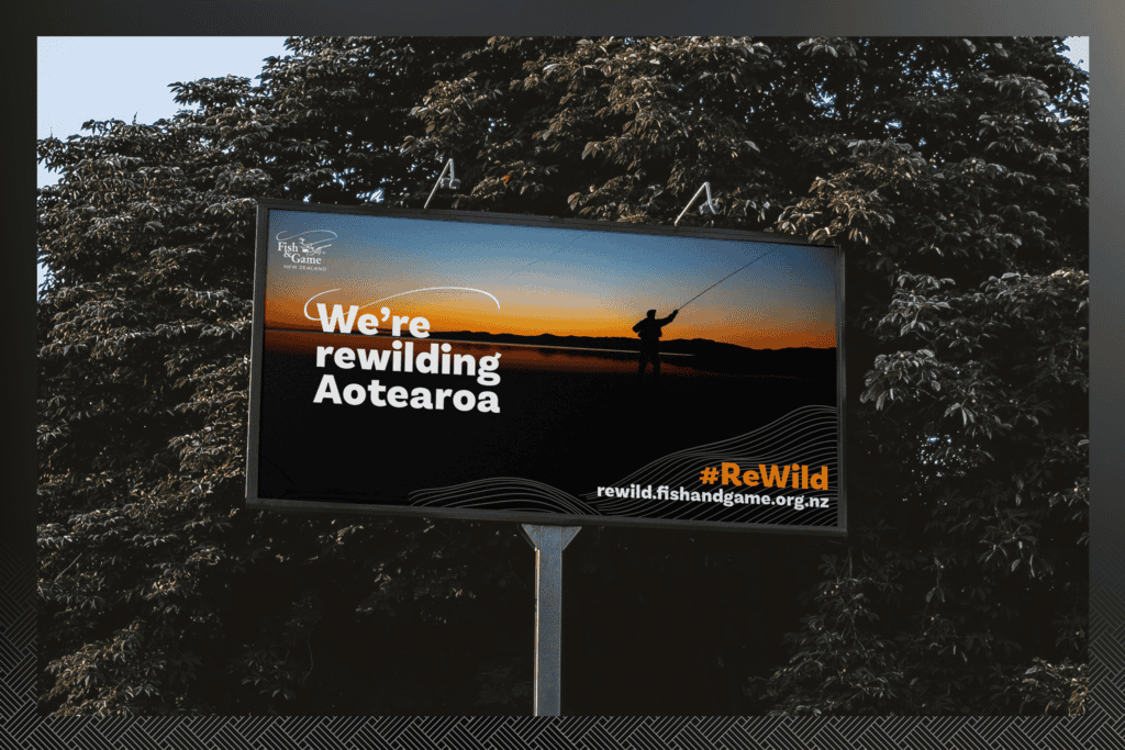 Fish-and-Game_ReWild-Portfolio-Mockups_Billboard-1 A billboard with the text "We're rewilding Aotearoa" and a web address rewild.fishandgame.org.nz, set against a background of trees.