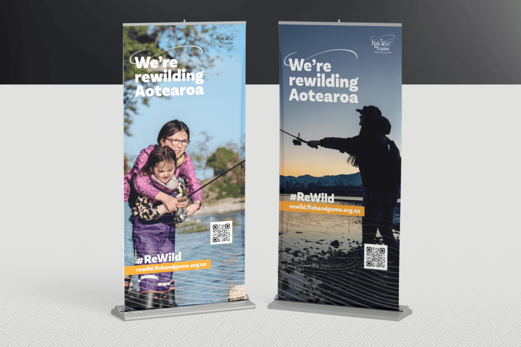 Fish-and-Game_ReWild-Portfolio-Mockups_Pull-up-banners Two vertical banners with the text "We're rewilding Aotearoa" display images of a child with a fishing rod and an adult fishing at sunrise. Both banners include a QR code and the hashtag #ReWild.