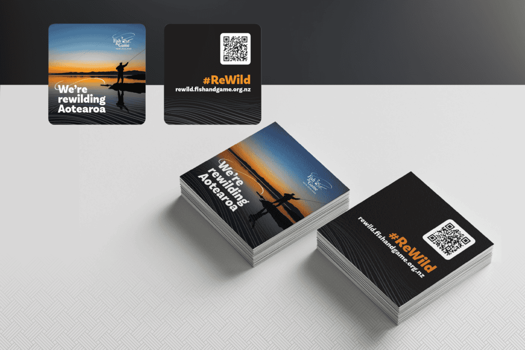 Fish-and-Game_ReWild-Portfolio-Mockups_QR-Card Two stacks of promotional cards on a textured surface. One stack features an image of a silhouette of a person fishing with the text "We're rewilding Aotearoa," and the other with #ReWild and a QR code.