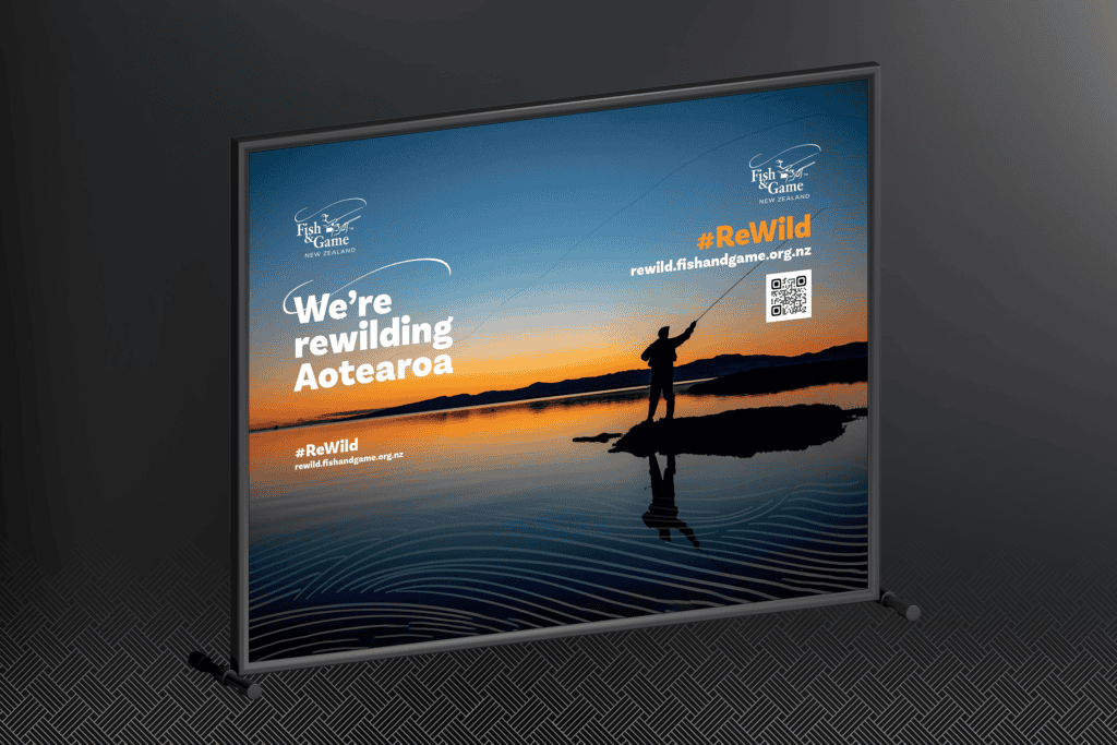 Fish-and-Game_ReWild-Portfolio-Mockups_selfie-wall Sign featuring a person fishing at a lake during sunset with the text "We're rewilding Aotearoa" and a website link: rewild.fishandgame.org.nz.