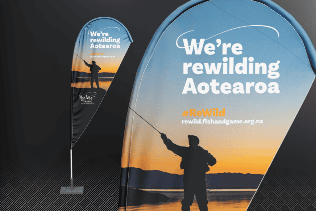 Fish-and-Game_ReWild-Portfolio-Mockups_teardrop-flag Banner promoting Fish & Game's rewilding campaign with the text "We're rewilding Aotearoa" and a website link. Image shows a person fishing at sunset.