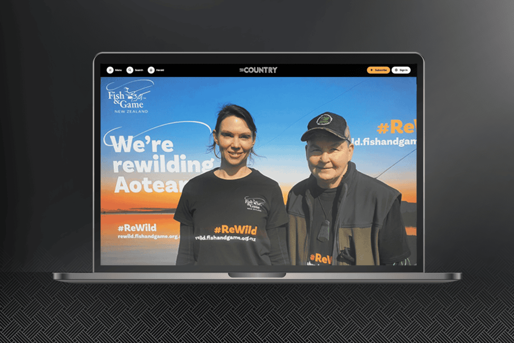 Fish-and-Game_ReWild-Portfolio-Mockups_Laptop_Dame_Lynda A laptop screen displaying a Fish & Game NZ website shows two people standing side by side and smiling. The website features a banner that reads "We're rewilding Aotearoa" along with the hashtags #ReWild. The people are wearing shirts with similar branding related to Fish and Game New Zealand.