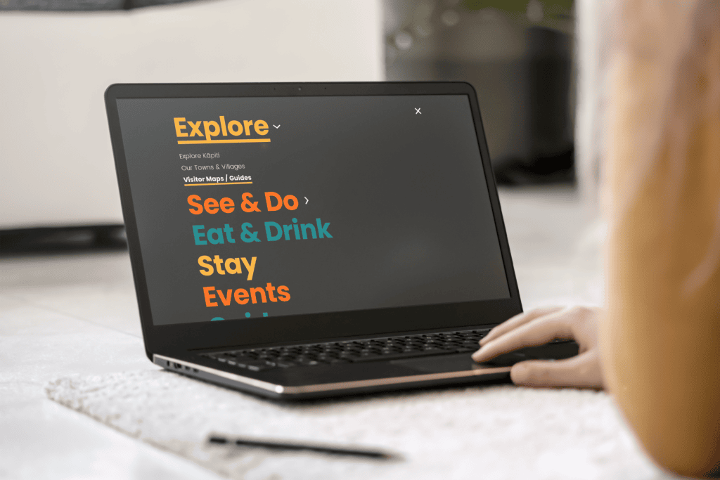 2_Visit-Kapiti_Portfolio-Mockup_1-0 A person using a laptop displaying a Kāpiti regional tourism website section titled "Explore" with categories: See & Do, Eat & Drink, Stay, Events. The text is brightly colored on a dark screen. The scene includes a carpet and a plant in the background.