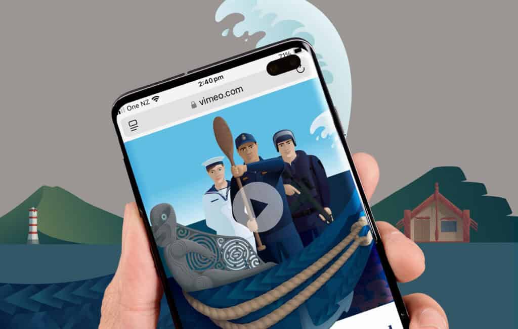 NZ-Navy-video-mobile A hand holding a smartphone displaying an illustration of three sailors in an animated video on vimeo.com. The background shows a large wave, a lighthouse, and green hills. A carved grey figurehead of a waka and rope are visible at the bottom of the phone screen.