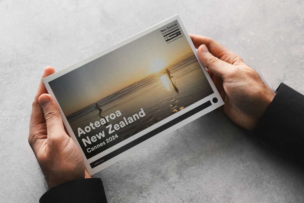 Cannes-booklet_1 A person holds a brochure featuring a beach scene with two people at sunset. The text reads "Aotearoa New Zealand Cannes 2024" with a New Zealand logo in the corner.