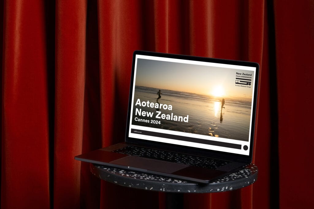 Cannes-laptop-redcurtain A laptop sits on a round table in front of red curtains. The screen displays an image of people walking along a beach at sunset with text reading "Aotearoa New Zealand Cannes 2024" and a logo for New Zealand Film Commission.