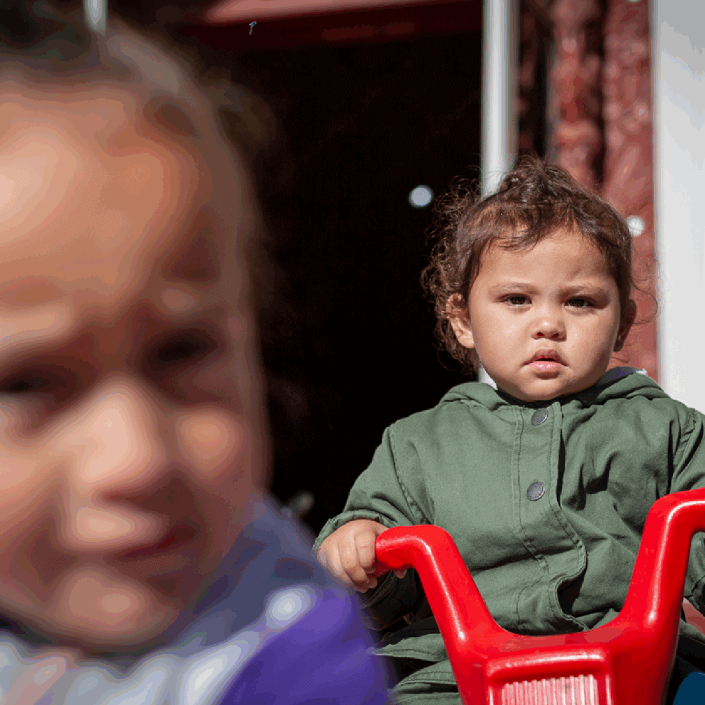 TToTW_PortfolioArtboard 1 copy 2 A young child in a green jacket sits on a red toy, looking at the camera, while another child in the foreground is out of focus. The scene appears to be outdoors near a building.