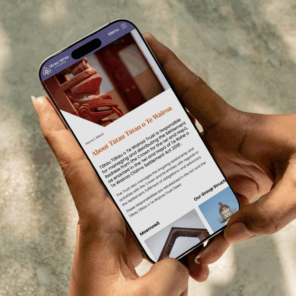 TToTW_PortfolioArtboard 1 copy 7 A person holds a smartphone displaying a webpage titled "About Tatau Tatau o Te Wairoa," featuring text and an image of a carved wooden figure, with sunlight illuminating their hands.