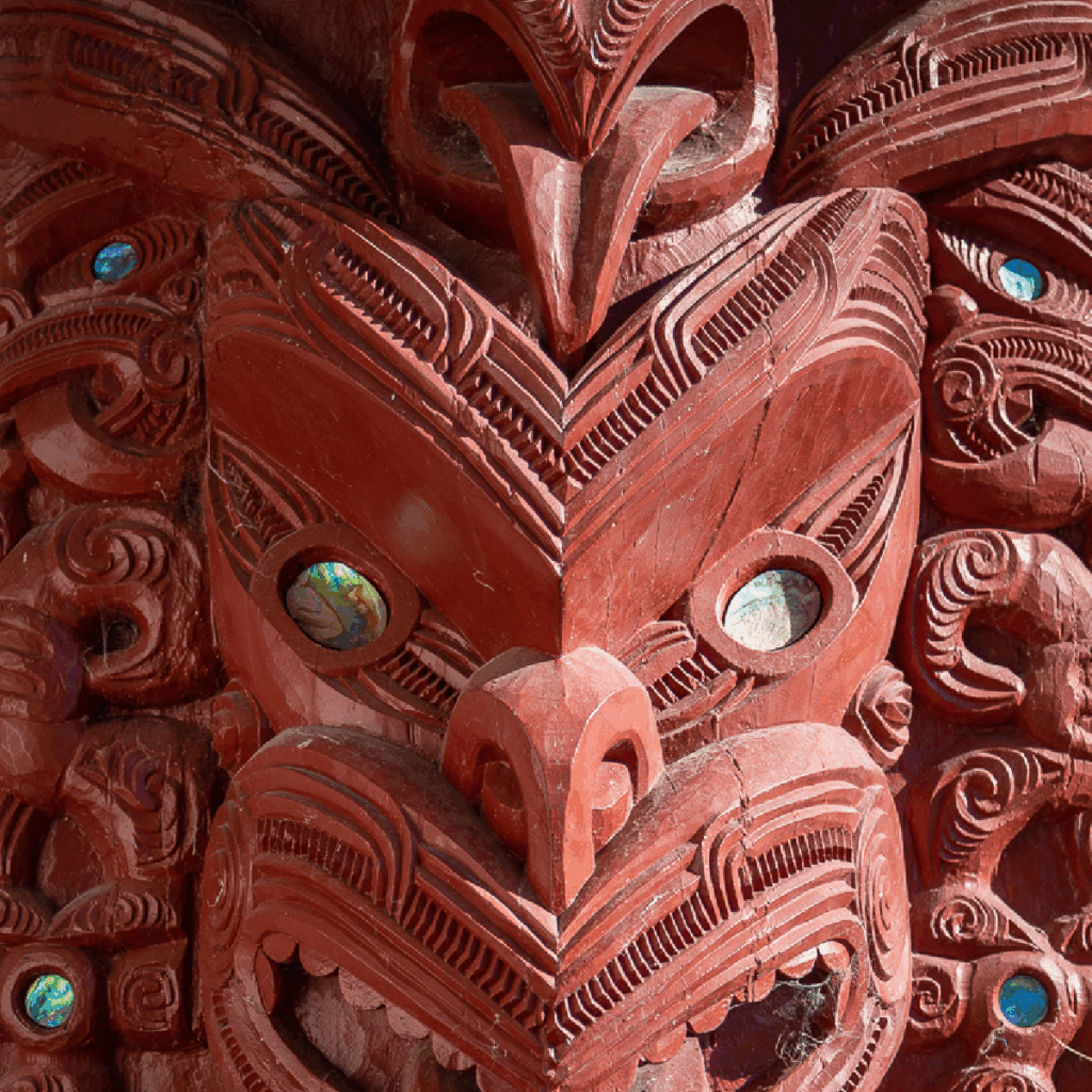 TToTW_PortfolioArtboard 1 copy 9 A close-up of a traditional Maori wooden carving features intricate patterns, inlaid paua shell eyes, and stylized facial expressions, showcasing rich red tones and elaborate detail.